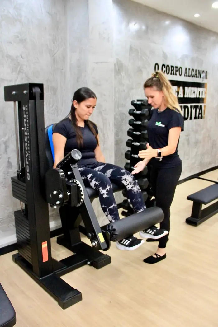 Professora orientando aluna em aparelho de musculação, com rack de halteres ao fundo