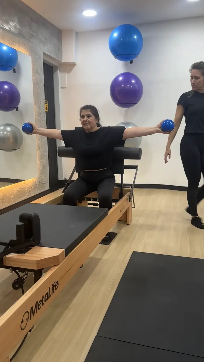 Aluna em pé no reformer segurando pesos, com professora ao lado corrigindo a postura